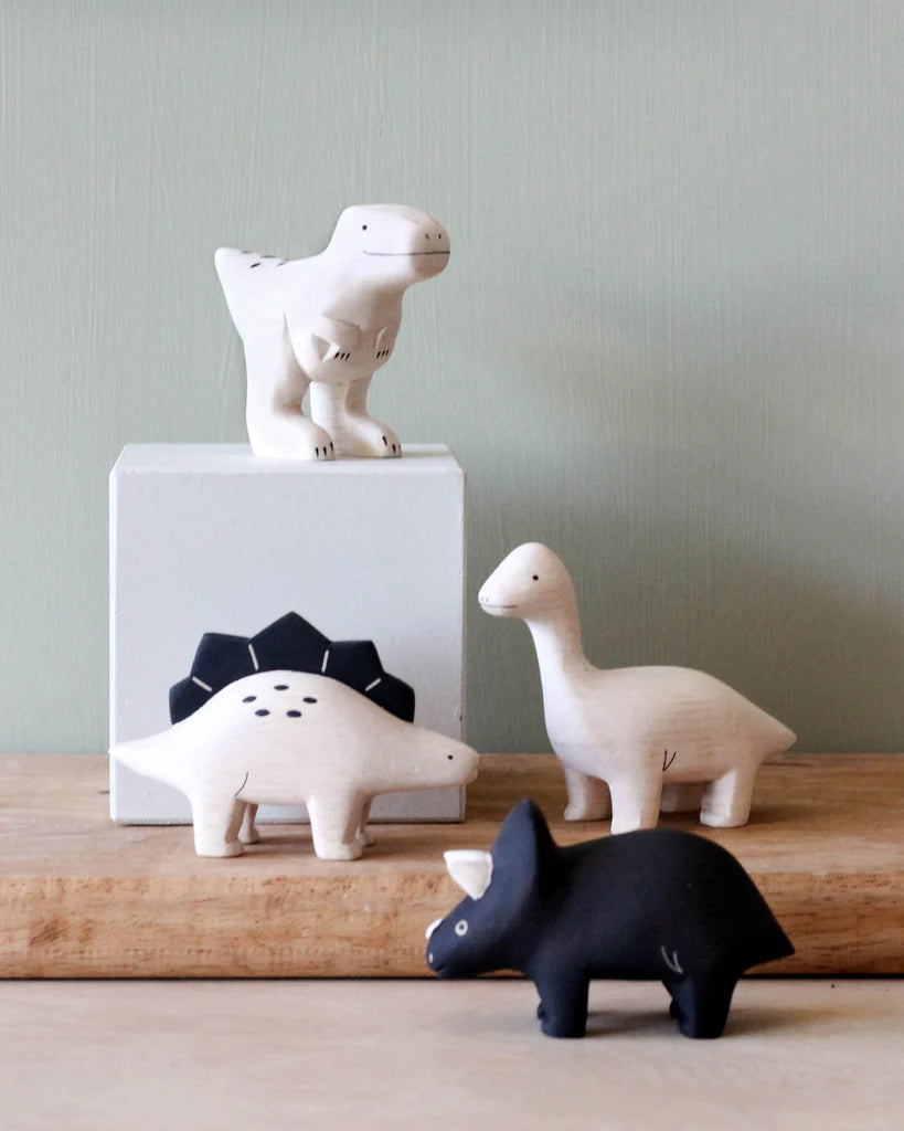 Four stylized dinosaur figurines handcrafted from Albizia wood displayed on a wooden surface against a green wall. The set includes a white T-rex on a white block, a Handmade Tiny Wooden Dinosaurs - Brachiosaurus.