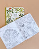 The Moulin Roty Botanist Coloring Book with Stockmar Pencil Set rests open on a tan surface, revealing black-and-white nature scenes inside—an ideal set for creative fun and imaginative coloring.