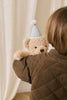 A young child with brown hair and a brown quilted jacket holds the Snuggle Bunnies - Birthday Arthur - Gingham plush toy, standing in front of a cream-colored curtain.