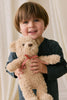 A young child with light brown hair and blue eyes smiles, holding Snuggle Bunnies' Arthur the Bear—Arthur with Red Stripe Bow—in front of a cream-colored curtain.