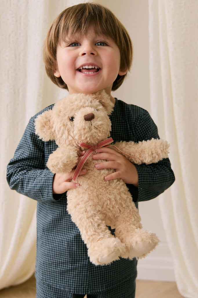 A young child with brown hair in checked pajamas smiles while holding Snuggle Bunnies' Arthur the Bear, featuring a red striped bow—the perfect children's gift. Light-colored curtains are visible in the background.