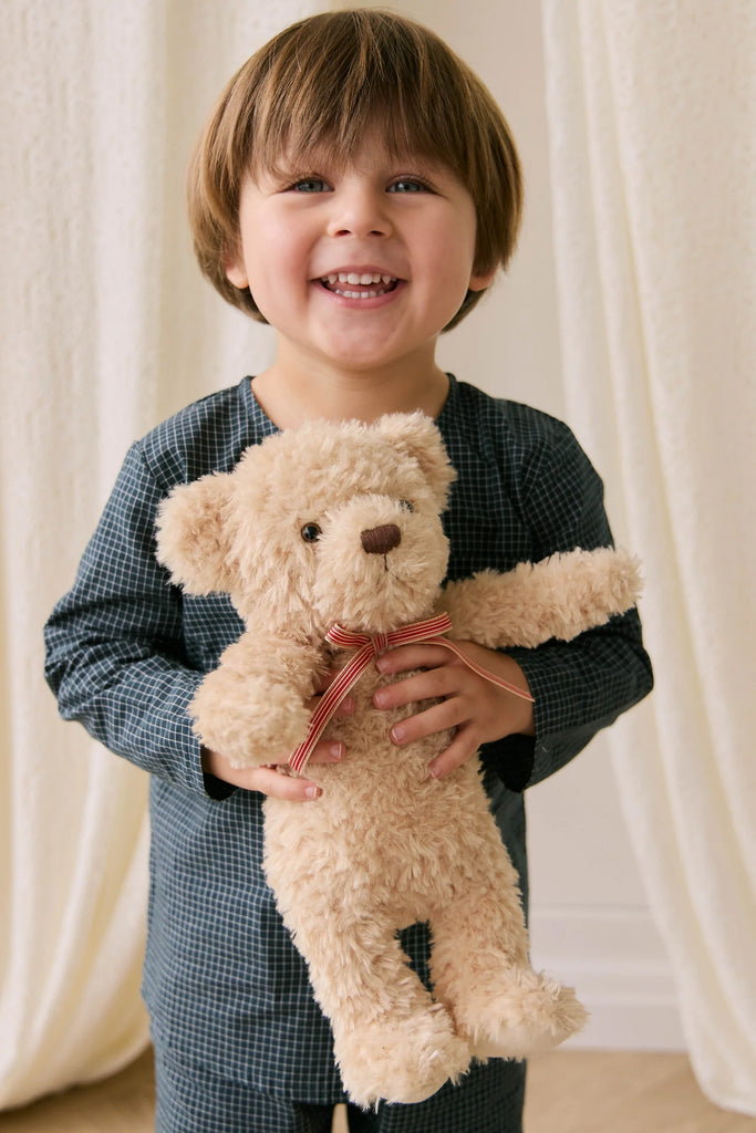 A young child with light brown hair and blue eyes smiles while holding Snuggle Bunnies—Arthur the Bear, featuring a red stripe bow, in front of cream-colored curtains—the perfect nursery companion.