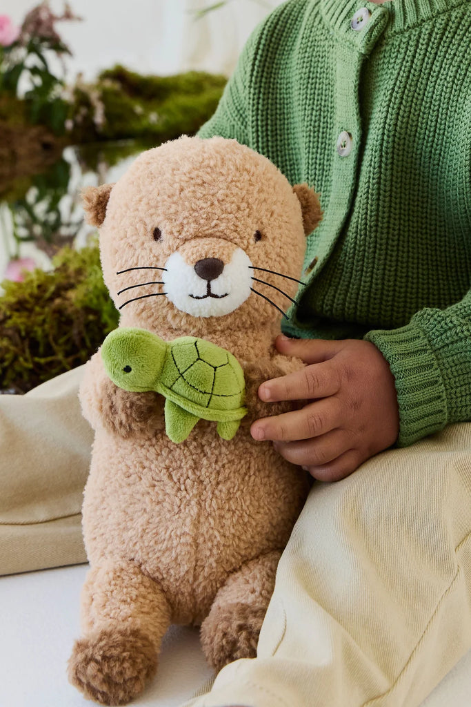 A tan Snuggle Bunnies - Ollie the Otter - Cocoa plush holding a small green turtle toy, held by a person wearing a green cardigan.