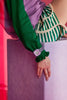 A person in a green long-sleeve top and striped green and white shorts wears the Mini Kyomo Watch - Lavender Field, sitting on a light purple block against a pink background with their hand resting relaxed on the edge.