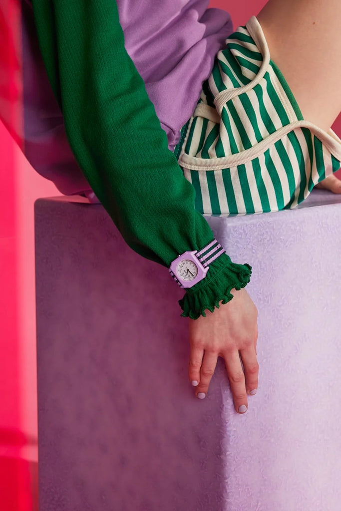 A person in a green long-sleeve top and striped green and white shorts wears the Mini Kyomo Watch - Lavender Field, sitting on a light purple block against a pink background with their hand resting relaxed on the edge.