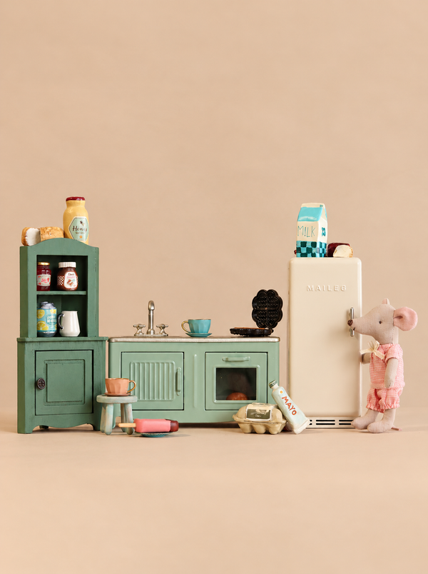 Maileg Little Sister’s Kitchen Set featuring a green hutch, cabinets, a cream refrigerator, a Maileg mouse in a pink outfit, play food, and dishware.