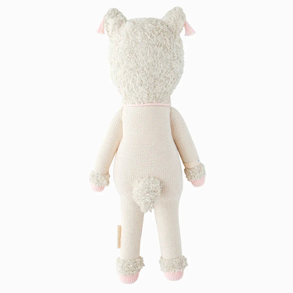 Cuddle + Kind Lola the Llama is a soft, light beige hand-knit doll shown from behind, featuring two pointed ears with pink tassels, a small fluffy tail, and pink accents on its cuffs and feet. Hypoallergenic and cuddly.