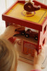A child plays with the Konges Sloejd Wooden Fire Station, designed in red and yellow from FSC-certified wood. They reach for a toy car on the second level while another sits on the roof's helipad, enjoying the detailed illustrations. Note: contains small parts that may be a choking hazard.