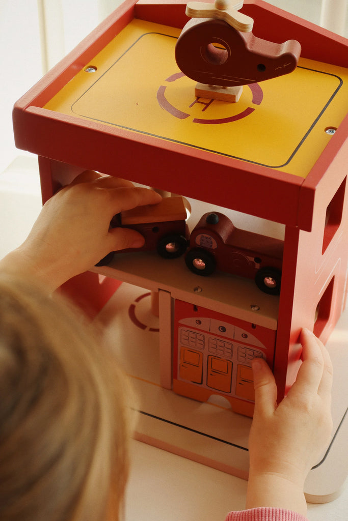 A child plays with the Konges Sloejd Wooden Fire Station, designed in red and yellow from FSC-certified wood. They reach for a toy car on the second level while another sits on the roof's helipad, enjoying the detailed illustrations. Note: contains small parts that may be a choking hazard.