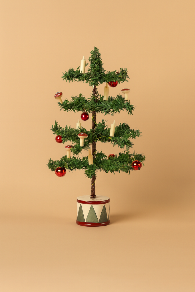 A Maileg Cozy Christmas Corner with Little Sister featuring a small artificial tree in a painted drum base, decorated with red ball ornaments, miniature candles, and mushroom ornaments.