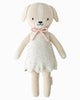 The Cuddle + Kind Mia the Dog is a beige hand-knit plush dog made from premium cotton yarn, dressed in a cream speckled dress with a pink bow at the neck, displayed on a plain white background.