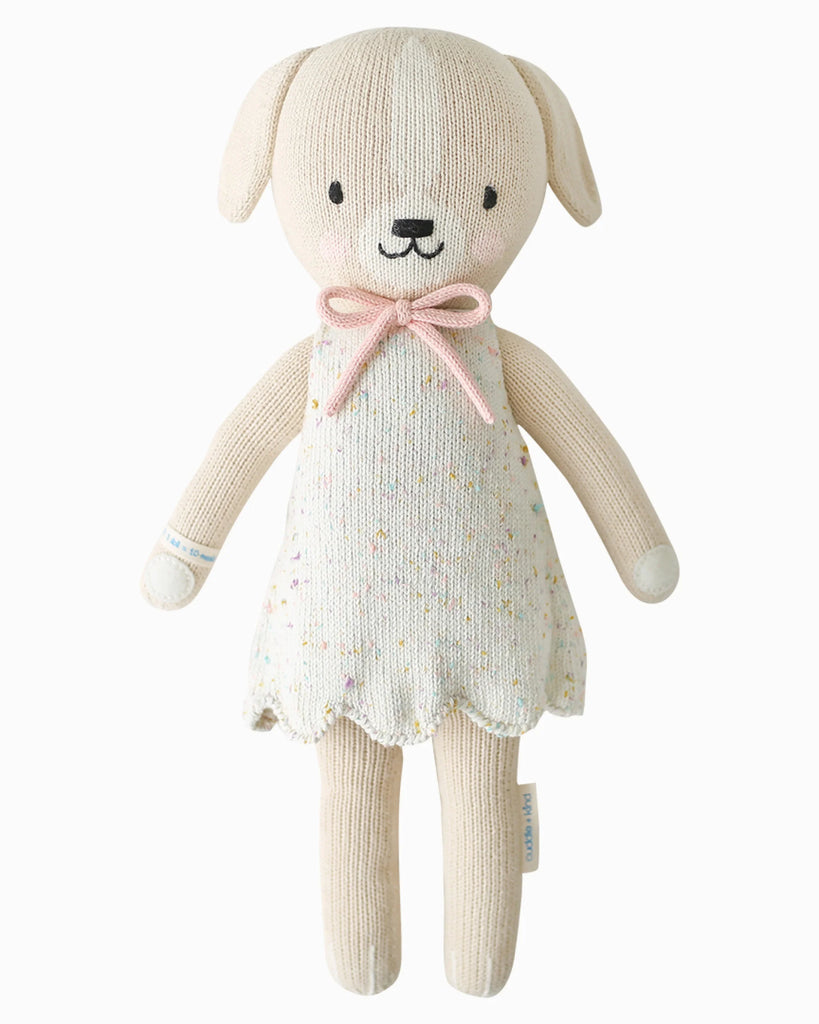 The Cuddle + Kind Mia the Dog is a beige hand-knit plush dog made from premium cotton yarn, dressed in a cream speckled dress with a pink bow at the neck, displayed on a plain white background.