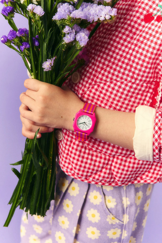 A person wearing a Mini Kyomo Watch - Ruby Papaya and a red checkered shirt holds a bouquet of purple flowers, paired with lavender pants featuring white daisy patterns.