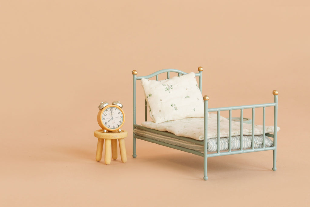 A small blue metal bed with floral bedding and a pillow sits next to the Maileg Stool Set, Mouse - Mint/Light Yellow, which holds a silver alarm clock, all set against a light beige background. The bed’s vintage paint finish adds extra charm to this cozy scene.