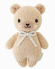 The Cuddle + Kind Baby Honey Bear in Robin’s Egg Blue, handmade in Peru from natural Peruvian cotton yarn, features black eyes, a small black nose, pink cheeks, and a light blue bow, set against a white background.
