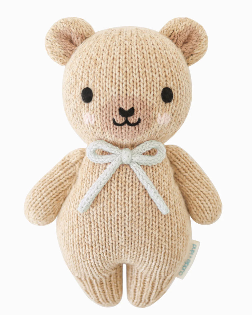 The Cuddle + Kind Baby Honey Bear in Robin’s Egg Blue, handmade in Peru from natural Peruvian cotton yarn, features black eyes, a small black nose, pink cheeks, and a light blue bow, set against a white background.