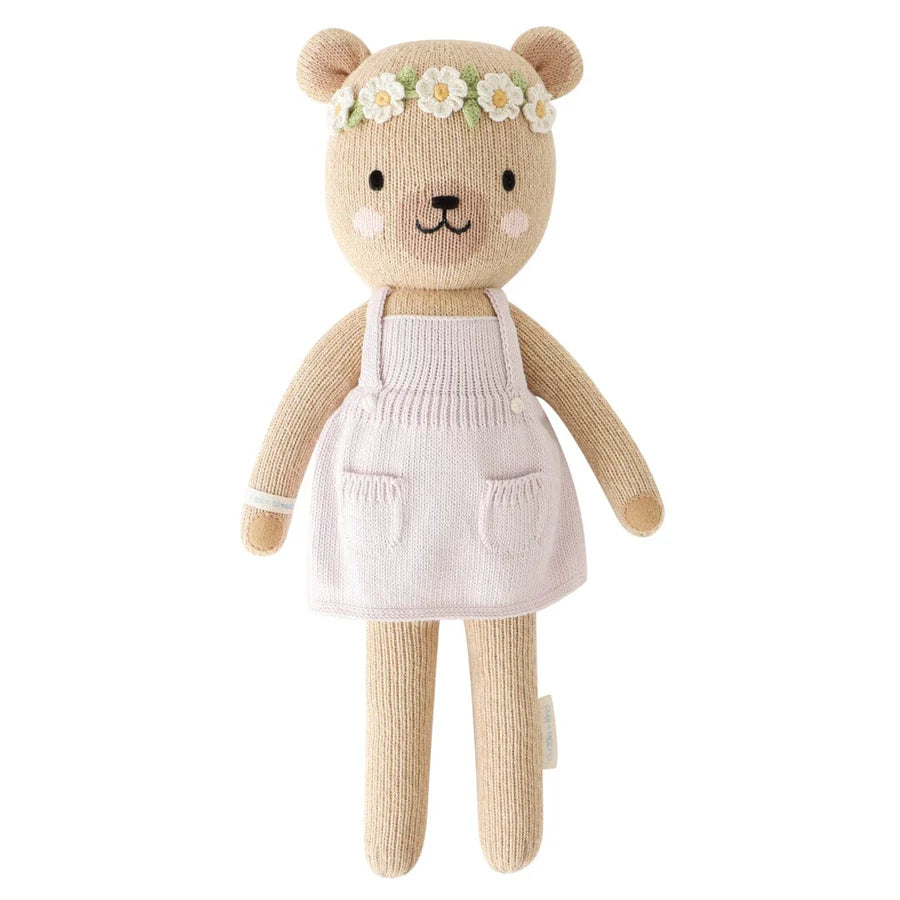 Cuddle + Kind Olivia The Honey Bear is a hand-knit teddy bear with light brown fur, a white dress, and a daisy flower crown. She has blushing cheeks and a happy expression, and is crafted using sustainable practices.