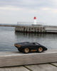 The Playforeer UFO Cannone Car in Black, a small toy with gold wheels inspired by 70s concept cars, rests on a wooden surface by the water, with a lighthouse and dock visible in the cloudy background.