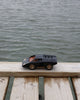 The Playforeer UFO Cannone Car in Black, inspired by 70's concept cars, sits on a wooden dock above the water with rippling waves in the background.