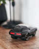 The Playforever Leadbelly Skeeter Car in Black, a small toy car with red tail lights, sits on a wooden surface. A blurred background shows a lamp and greenery.