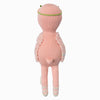 Cuddle + Kind Penelope the Flamingo is a pink, hand-knit doll with a round head, small tail, green headband, fluffy vest, and white crisscross ribbons on its feet. The doll stands upright facing away.