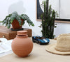 A wooden desk displays a round terracotta vase, straw hat, Playforever MINI KEN CASH Blue racing car, potted plants, stack of books, and a framed picture against a white wall.