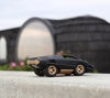 The Playforeer UFO Cannone Car in Black, featuring a sleek geometric design inspired by 70’s concept cars, sits on smooth concrete. A modern round building with a dark wood exterior is blurred in the background.