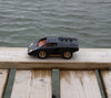 The Playforeer UFO Cannone Car in Black, featuring tan windows and wheels, sits on a wooden dock above rippling water, evoking 70’s concept car style.