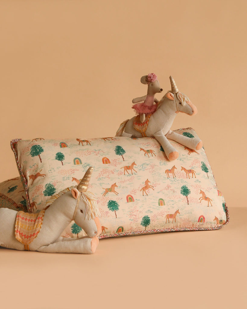 Two plush unicorn toys and a small mouse with a hat are arranged on and beside the Maileg Cushion - Unicorn, a pastel rectangular cushion made of soft cotton with unicorn and rainbow patterns, set against a gentle beige background.