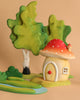 The Bumbu Wooden Toadstool House features a charming, round mushroom home with a red, white-spotted roof, surrounded by green trees, shrubs, and a blue stream on a beige background—ideal for imaginative play.