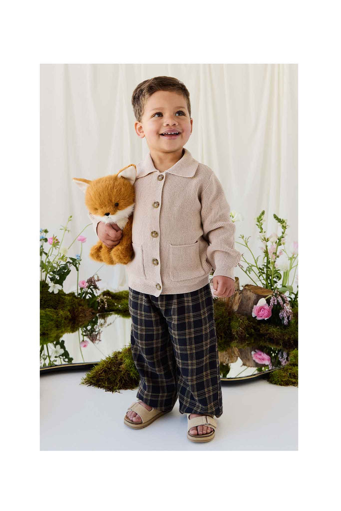 A young child wearing a beige cardigan and plaid trousers smiles while holding a Snuggle Bunnies - Archie the Fox - Natural plush.