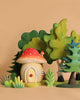 The Handmade Wooden Mushroom House features a red and white spotted roof and sits among green trees and bushes, creating a Montessori and Waldorf-inspired scene on a plain beige background.