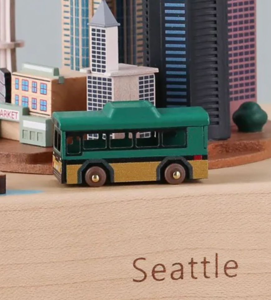 The Replacement Bus For Seattle Music Box showcases a green bus prominently in the foreground, symbolizing efficient bus replacement. Behind it, Seattle's skyline and the iconic Space Needle add to the scene, while "Seattle" is elegantly engraved on the base.