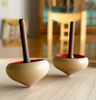 Two Mader Zwirbel Spinning Tops - Green, crafted from Walnut and Ash with dark handles and red interiors, rest on a smooth light-colored surface. A wooden chair and cabinet appear softly blurred in the background.