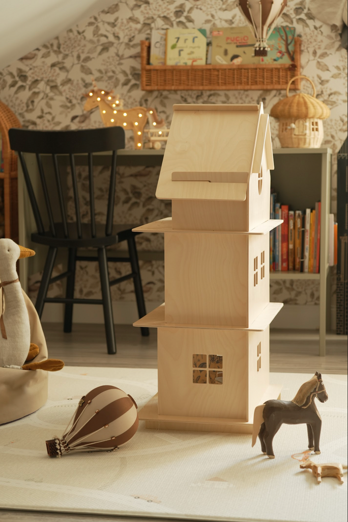 A cozy children's room features a wooden dollhouse, toy horse, and a handmade Hot Air Balloon - Light Brown / Dark Brown on a light rug. In the background are a black chair, desk, books, and soft patterned wallpaper for charming decor.
