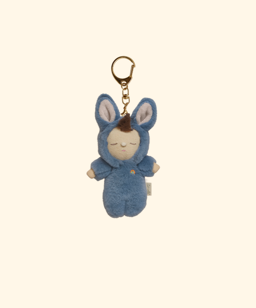 The Olli Ella Cozy Bag Charm - Bunny Twiggy is a plush bag charm featuring a small doll in a blue bunny costume with large ears, closed eyes, and a peaceful expression, attached to a gold keyring on a plain beige background.
