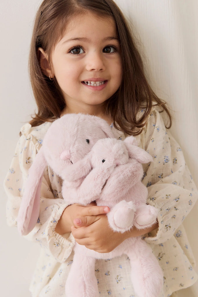 A young girl with brown hair smiles as she hugs her Snuggle Bunnies - Frankie the Hugging Bunny - Blossom, cuddling the super soft plush to her chest while wearing a cream floral long-sleeve shirt.