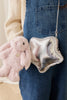 A person in denim overalls holds a plush pink bunny and wears a shiny silver star-shaped purse adorned with the Snuggle Bunnies - Penelope Keyring - Blossom on the shoulder strap.