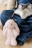 The Snuggle Bunnies - Penelope Keyring - Blossom, a plush pink bunny bag charm with long ears, is clipped to a shiny silver bag resting on someone's lap. The person wears a beige top and blue denim overalls.