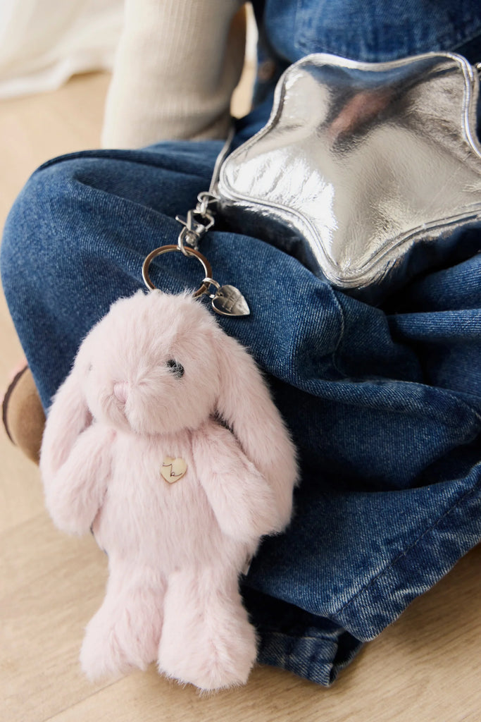 The Snuggle Bunnies - Penelope Keyring - Blossom, a plush pink bunny bag charm with long ears, is clipped to a shiny silver bag resting on someone's lap. The person wears a beige top and blue denim overalls.