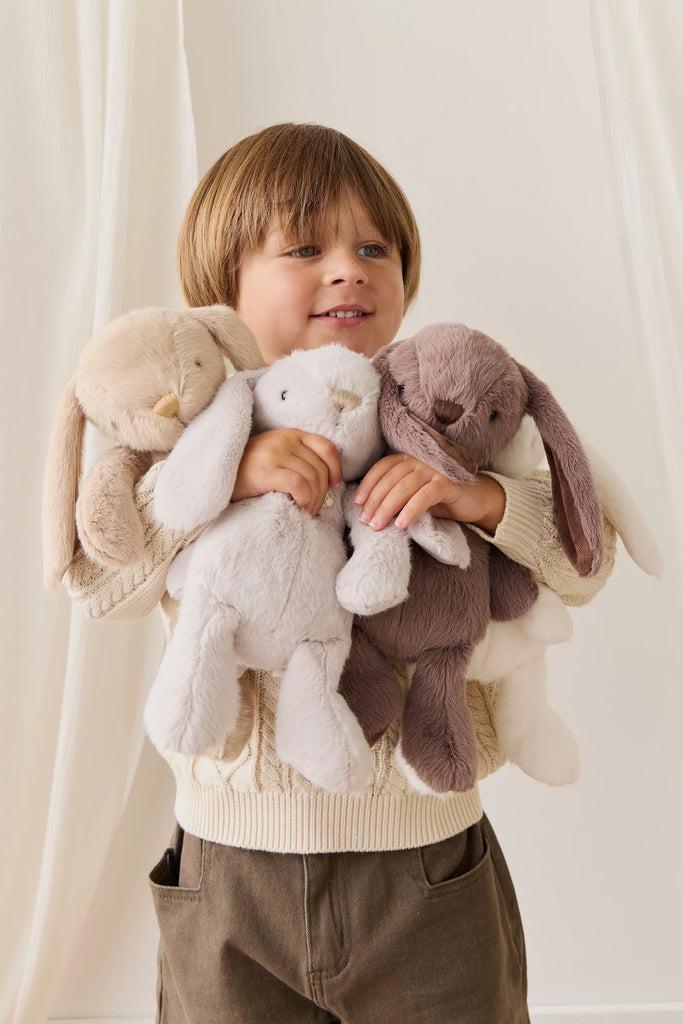 A young child smiles while hugging Snuggle Bunnies - Penelope - Caramel, a soft plush bunny toy, against a light, gentle background.