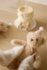 A person in white mittens holds the Snuggle Bunnies - Birthday Arthur - Fairy plush toy with a pink party hat in front of a small frosted cake topped with mini teddy bears. Another keepsake Snuggle Bunnies plush is nearby on the light wooden surface.