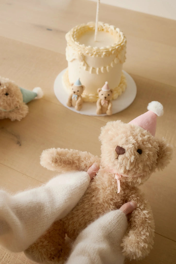 A person in white mittens holds the Snuggle Bunnies - Birthday Arthur - Fairy plush toy with a pink party hat in front of a small frosted cake topped with mini teddy bears. Another keepsake Snuggle Bunnies plush is nearby on the light wooden surface.
