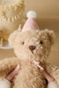 Two hands hold Snuggle Bunnies - Birthday Arthur - Fairy, a fluffy beige bear plush in a pink party hat with white pom-pom. Behind it, a decorated cake and soft lighting set the scene for this adorable keepsake gift.