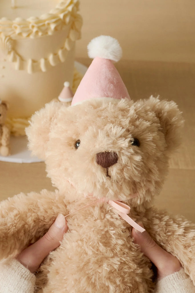 Two hands hold Snuggle Bunnies - Birthday Arthur - Fairy, a fluffy beige bear plush in a pink party hat with white pom-pom. Behind it, a decorated cake and soft lighting set the scene for this adorable keepsake gift.