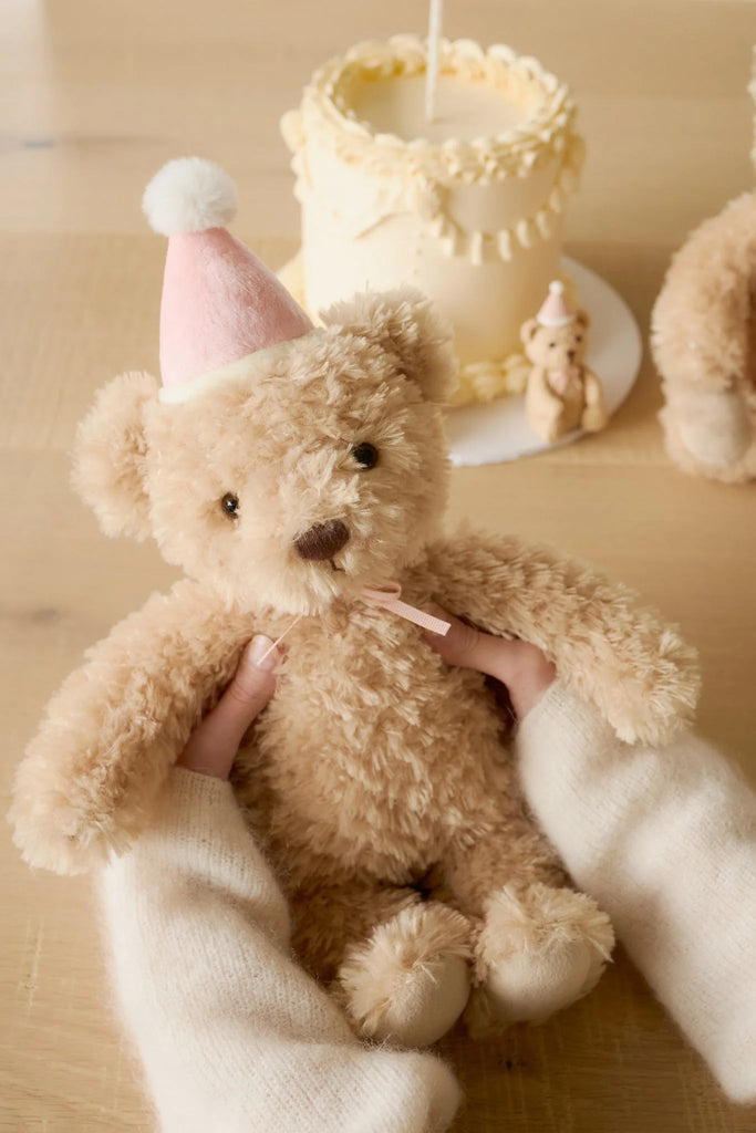 A person with cream-colored sleeves holds the Snuggle Bunnies - Birthday Arthur - Fairy plush, featuring a pink party hat, with a decorated cake and another teddy bear in the background on a light wooden table.