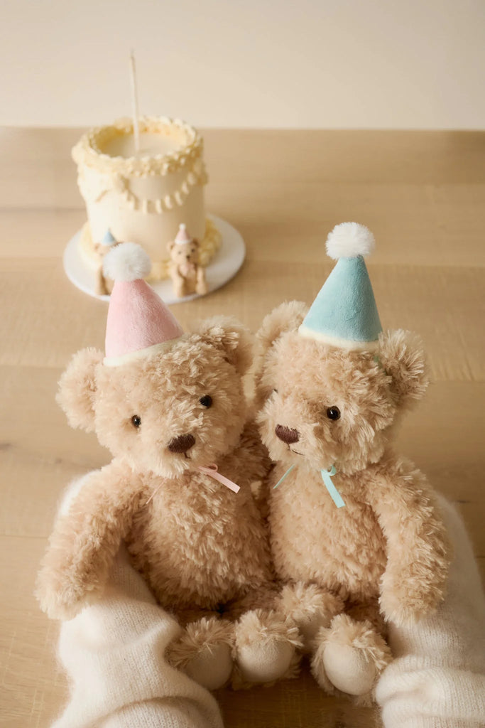 Two plush Snuggle Bunnies – Birthday Arthur and Fairy – wear pastel party hats as they're held together by gloved hands. A birthday cake with a candle sits behind them, making this the perfect keepsake gift for any special birthday bunny.