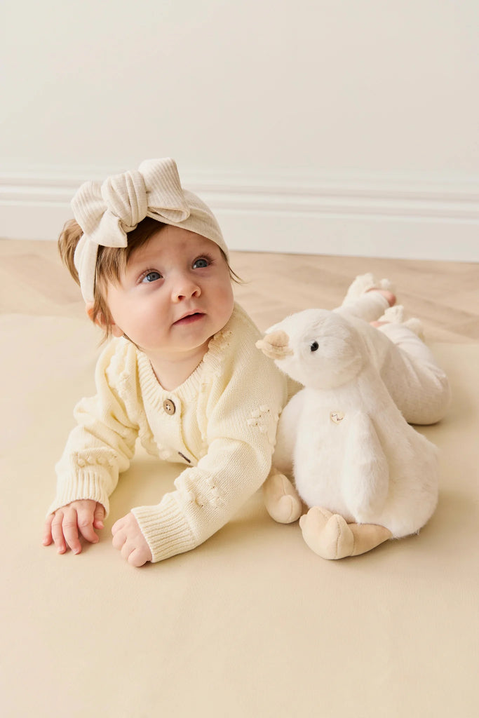 A baby in a cream knit sweater with a big matching headband lies on their stomach next to a super soft Snuggle Bunnies - Rose The Duck plush toy on a light-colored floor.