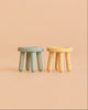 Two small Maileg Stool Set, Mouse stools—one in mint and one in light yellow—crafted from beech wood with a vintage paint finish, sit side by side on a peach-colored surface against a matching peach background.