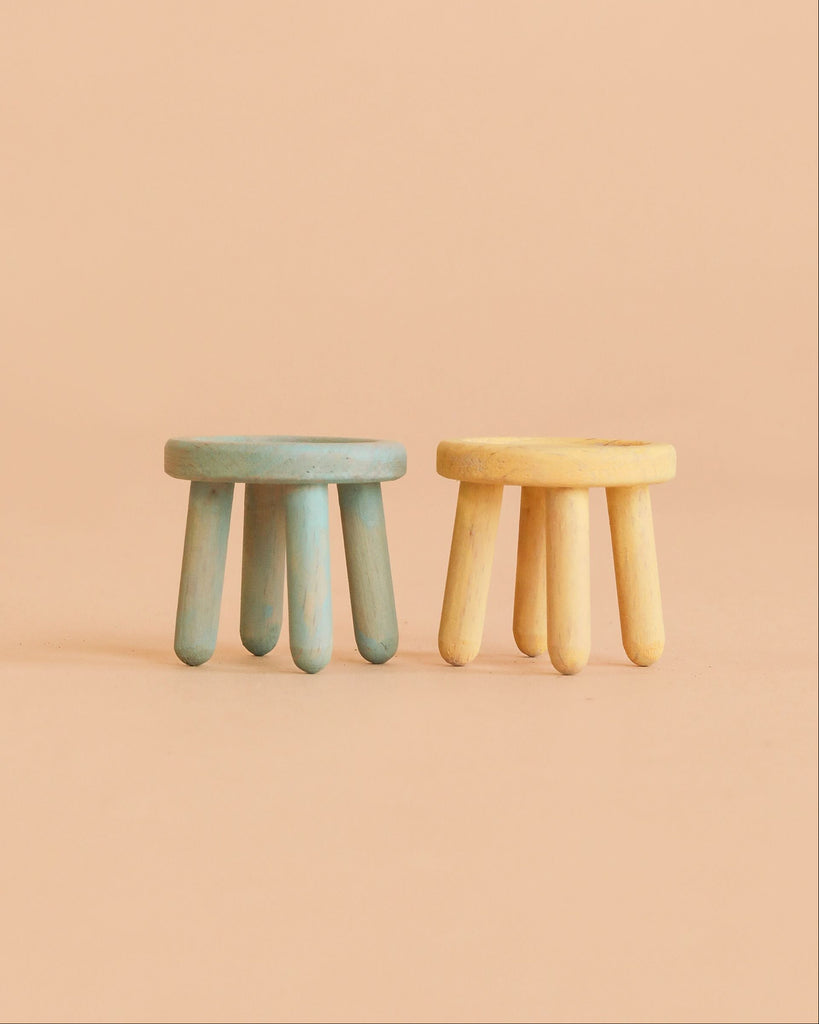 Two small, round wooden stools with thick, cylindrical legs—one painted blue and the other yellow—are placed side by side on a plain peach-colored background. Part of the Maileg Family Room Set, these stools make perfect Maileg mouse furniture or charming dollhouse accessories, adding a delightful touch to any miniature Maileg scene.